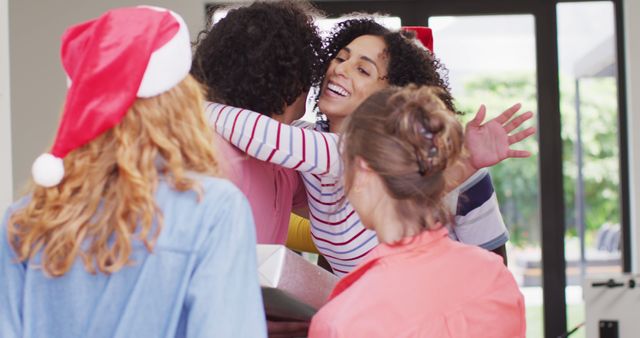 Group of friends gathering indoors for a joyful holiday celebration. They are exchanging warm embraces and dressed in festive holiday attire, including Santa hats. This image is perfect for use in advertisements promoting holiday events, parties, and greeting card designs.