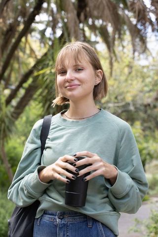 Young woman holding travel mug on park path wearing green sweatshirt, casual outdoor