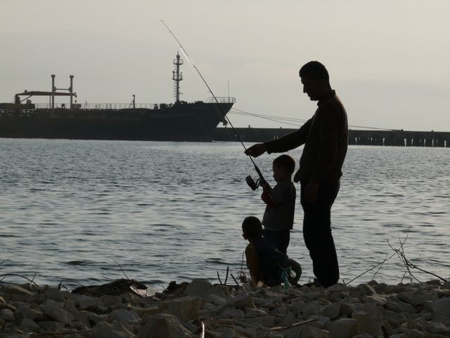 Silhouette of father teaching young children fishing beside rocky beach with dock and cargo ship close by. Perfect for showcasing family togetherness, bonding and outdoor activities. Suitable for campaigns centered on fatherhood, patience, outdoor skills, leisure activities, family time.