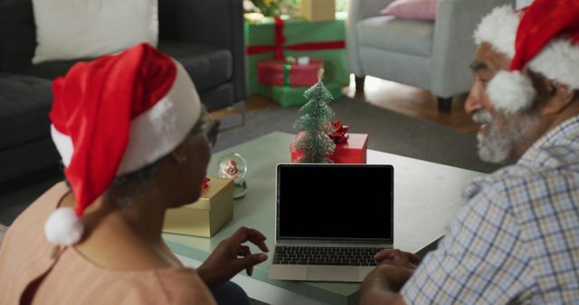 Two seniors are using a laptop for a video call during a Christmas celebration. They are wearing Santa hats and are surrounded by festive holiday decorations, including a small Christmas tree and wrapped gifts. This image is suitable for holiday greeting card designs, promoting virtual holiday gatherings, or illustrating family connections during Christmas.