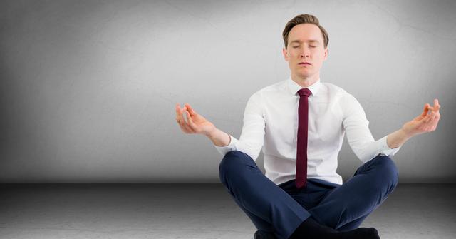 Businessman in formal attire meditating with eyes closed, seated in lotus position against empty grey wall. Ideal for concepts of corporate wellness, stress relief, and mindfulness practices in workplace settings. Can be used in brochures, blog posts, or websites promoting mental health and wellness programs in corporate environments.