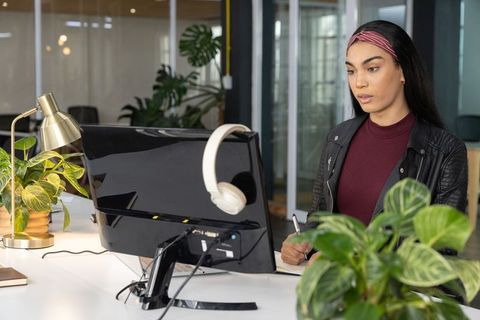 Young professional woman working at modern office desk with monitor, notebook and plants