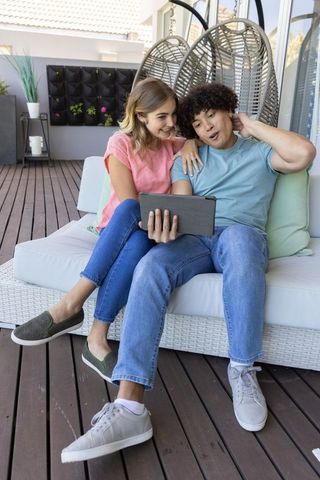 Young couple relaxing on woven outdoor sofa sharing tablet on sunny wooden patio