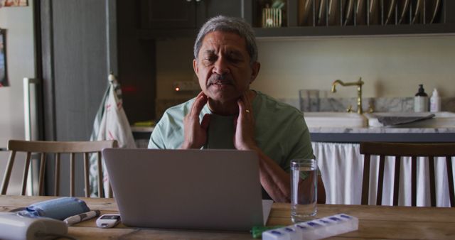 Elderly man sitting in kitchen using laptop for virtual healthcare consultation. Hands on neck indicating potentially a sore throat. Scene captures essence of telemedicine and online health consultations. Ideal for illustrating remote healthcare, telehealth services, senior digital engagement, virtual medical advice, home health monitoring and self-care awareness.