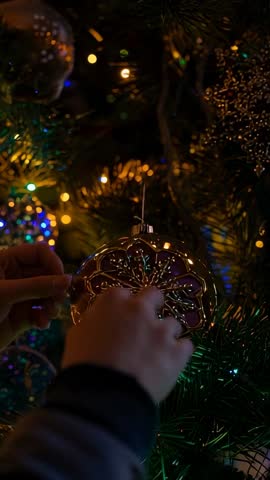 Vertical video Hanging gold filigree ornament on lit Christmas tree by dark-sleeved hands