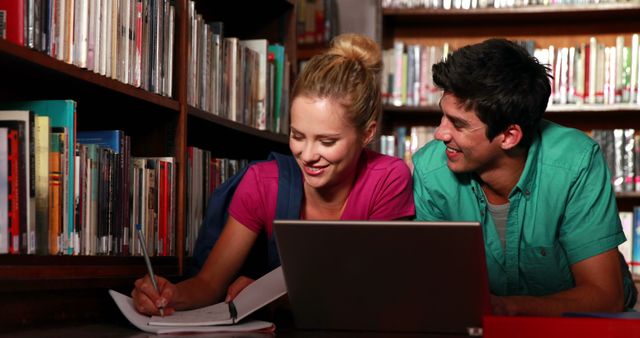 Two students are smiling while studying together in a library. They are using a laptop and taking notes in notebooks surrounded by bookshelves. Perfect for illustrating themes of education, teamwork, and collaborative learning in educational materials, websites, or promotional content for academic institutions.