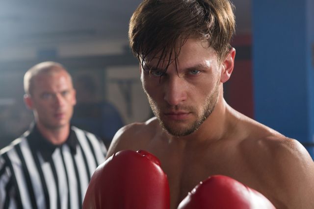 Young male boxer wearing red gloves standing in boxing ring with intense expression, referee in background. Ideal for use in sports-related content, fitness promotions, motivational materials, and advertisements for boxing gyms or training programs.