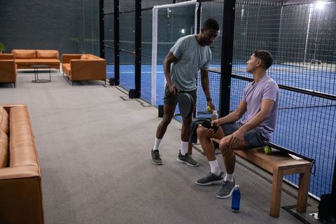Mixed race friends chatting beside indoor padel court after match holding paddle and phone