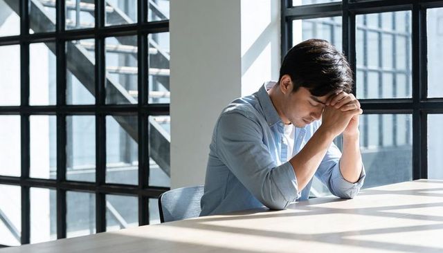 Man leaning forward holding head at desk in sunlit industrial office, minimalist workspace