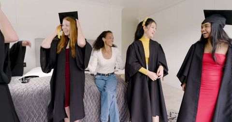 Diverse graduates adjusting mortarboards and celebrating on bed in bedroom