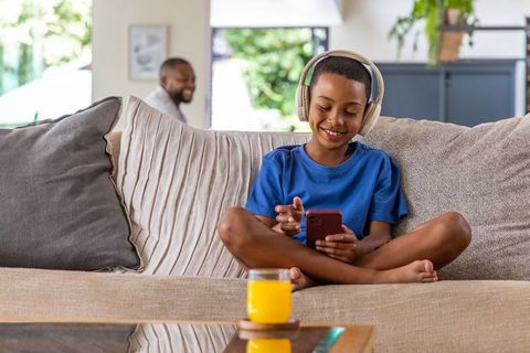 African american boy wearing headphones relaxing on couch with smartphone and orange juice