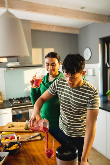 Multiracial young man looking at boyfriend pouring smoothie in glass on kitchen island, copy space. Drink, healthy, unaltered, love, togetherness, homosexual, lifestyle and home concept.