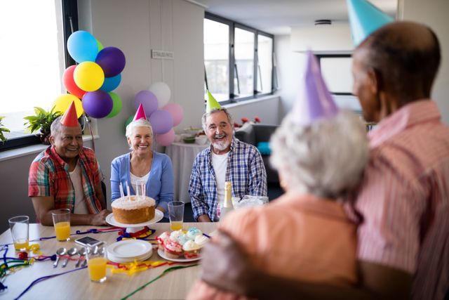 Senior friends celebrating a birthday party with cake and colorful balloons. They are wearing party hats and smiling, creating a joyful atmosphere. This image can be used for promoting senior living communities, birthday party invitations, or advertisements focusing on elderly social gatherings and celebrations.