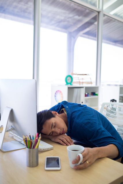 Male executive resting his head on desk in office, holding coffee cup, with computer and smartphone nearby. Ideal for illustrating workplace fatigue, stress, overwork, need for breaks, and work-life balance.
