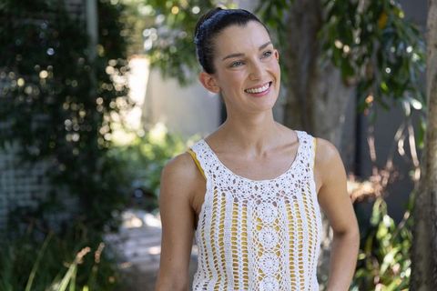 Smiling woman standing in shaded backyard garden wearing white crochet top over yellow tank