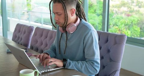 Man working remotely at dining table typing on laptop wearing pink headphones in modern home