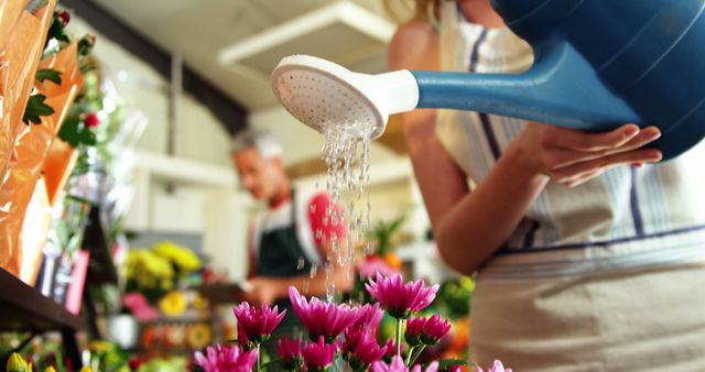 Florist using blue watering can to water vibrant pink flowers in a flower shop. Great for florist advertisements, gardening blogs, and articles focused on plant care, horticulture, and small business operations. The image evokes themes of nurture, growth, and the everyday tasks within a florist shop.