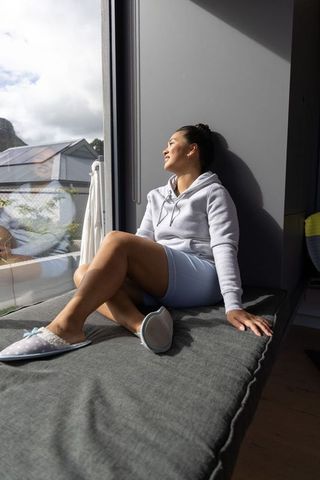 Young woman relaxing on sunlit window seat gazing outside cozy minimalist home interior