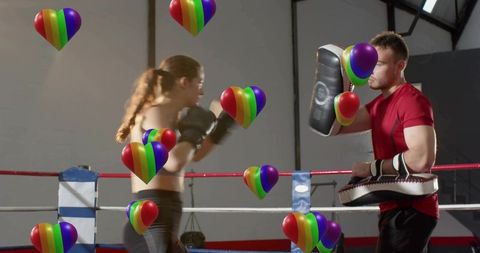 Female boxer training with coach in ring, punching mitts amid colorful rainbow hearts