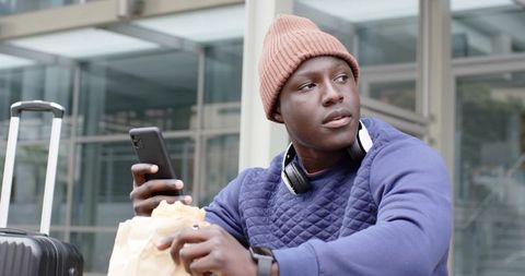 African American traveler checking phone while waiting with carry-on luggage and snack