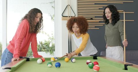 Diverse female friends playing pool together in modern sunlit home lounge