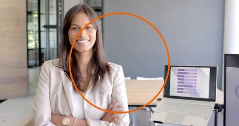 Confident businesswoman standing with arms crossed smiling in modern office with laptop chart