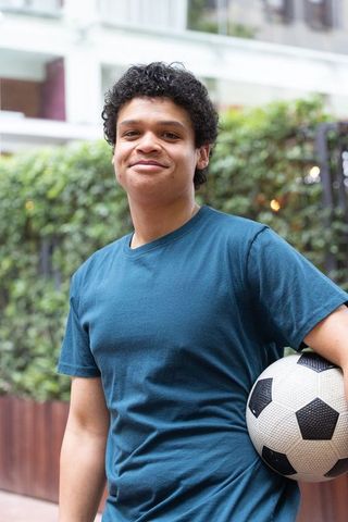 African american teen holding soccer ball smiling in urban courtyard with greenery
