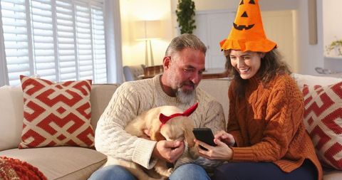 Middle-aged couple celebrating Halloween at home with dog wearing horns and pumpkin hat