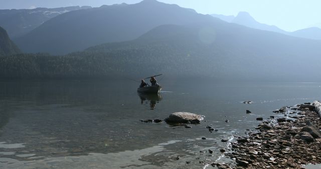 A small boat with two people is gently floating on a still mountain lake. The tranquil water and the surrounding landscape of distant ridge-lined mountains and mist create a serene atmosphere. Perfect for uses involving outdoor activities, nature retreats, tranquility themes, adventure marketing, or environmental conservation campaigns.
