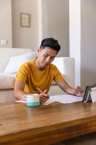 Asian man working at home writing documents and checking tablet on coffee table