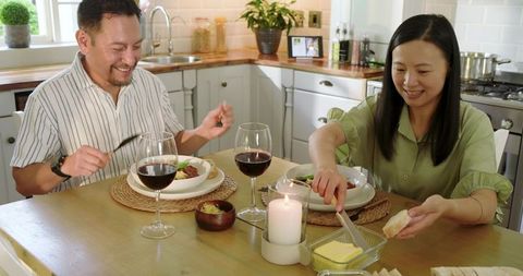 Asian couple sharing candlelit dinner, spreading butter and smiling over wine and bread