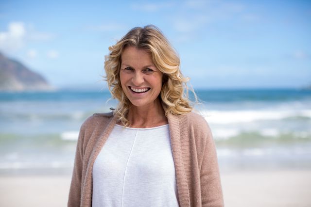 Mature woman with blonde hair smiling while standing on the beach with ocean waves in the background. Ideal for use in lifestyle, travel, and wellness content, promoting relaxation, happiness, and coastal living.