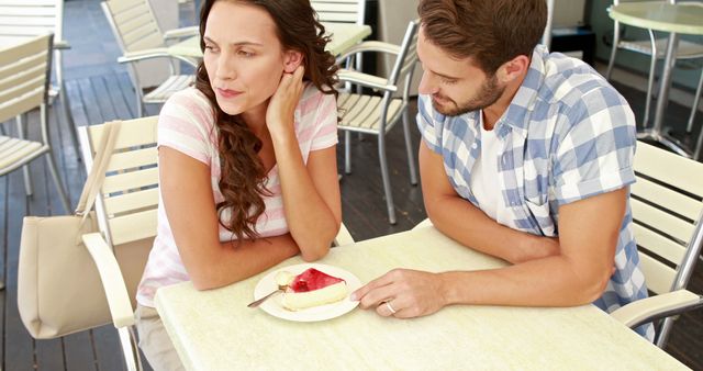 Couple sitting at a table outdoors with a dessert plate. Both individuals are looking away, suggesting a moment of tension. Suitable for concepts related to relationships, communication, and emotional moments. Also useful for lifestyle and cafe settings.