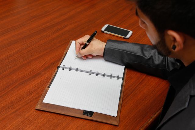 Business professional writing in notebook at wooden desk, smartphone to the side. Ideal for business, productivity, office environments, and professional contexts.