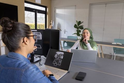 Muslim woman and colleague reviewing architectural floorplans on laptops in modern office