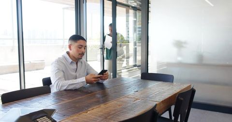 Businessman checking smartphone in conference room with rustic wooden table and glass doors