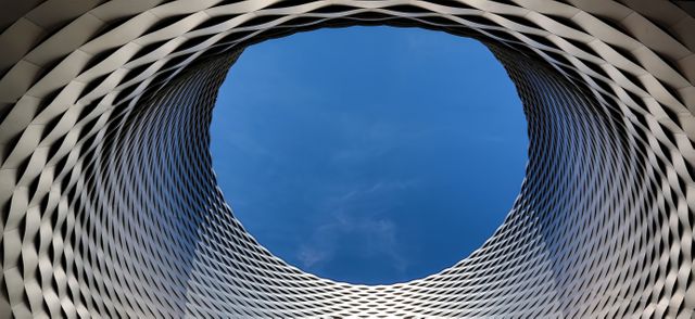 This stock photo features a modern architectural structure with a circular opening that reveals a blue sky. The geometric, metallic design creates a sense of symmetry and innovation. This image is suitable for use in publications related to architecture, urban development, modern design trends, or as an abstract background. It can also be used to convey themes of creativity, innovation, and forward-thinking.
