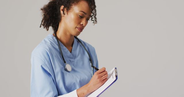 Depicts a confident female nurse in a blue uniform and stethoscope writing on a clipboard. Perfect for healthcare-related content, medical brochures, educational materials, hospital website graphics, and professional medical presentations. Highlights dedication and professionalism in a medical setting.