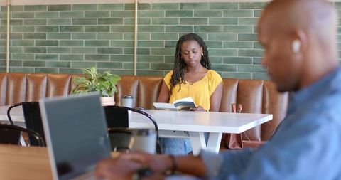 Woman reading paperback in cozy cafe coworking space with laptop, plant and tiled wall