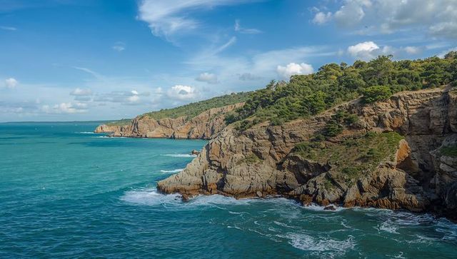 Turquoise coastal cliffs with pine-covered headland and crashing waves under blue sky