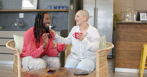 Friends laughing over coffee in cozy kitchen lounge, man with dreadlocks and bald woman