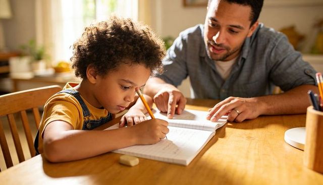 Father helping child with homework at sunlit kitchen table, writing in notebook