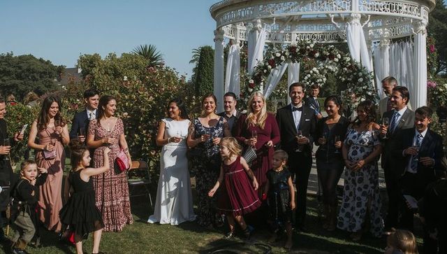 Garden wedding guests celebrating by floral gazebo with bride in white gown and groom