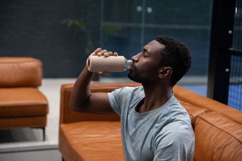 African american man drinking from reusable bottle on leather sofa in modern lounge