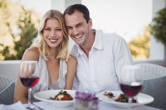 Romantic couple enjoying a meal together at an outdoor restaurant. Both are smiling and appear happy, with wine glasses and plates of food in front of them. Ideal for use in advertisements for restaurants, dating services, lifestyle blogs, and romantic getaway promotions.