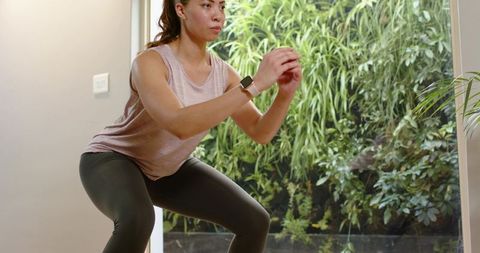 Woman performing squat workout at home near glass door with green garden view and smartwatch