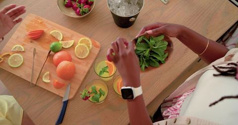 Friends preparing citrus mocktails with fresh mint, strawberries and ice on wood table