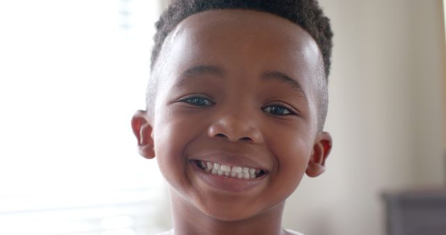 Cheerful young African American boy with a bright smile and natural hair indoors. Perfect for usage in family-oriented themes, children's services, educational materials, advertisements targeting parents, or magazines focused on childhood joy and happiness.