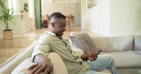 Smiling african american man relaxing on light sofa and using smartphone in modern home