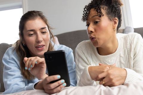 Diverse female friends lounging on couch sharing smartphone and laughing in cozy room